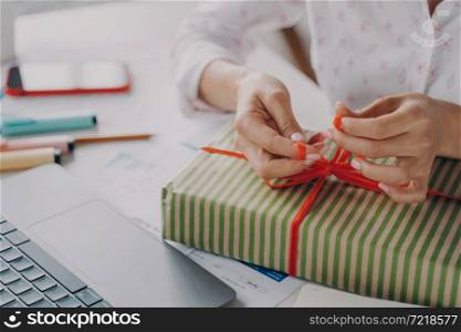 Cropped shot of woman online store business owner packing package gift box for customer during Christmas winter holidays, female tying red bow on xmas present, preparing parcel on table. Female hands packing package gift box for customer during Christmas winter holidays