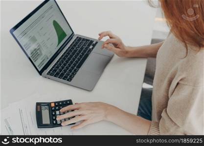 Cropped shot of woman using laptop and calculator while sitting at table at home, looking at computer screen with diagrams, calculating bills or household expenses, female accountant working remotely. Woman using laptop and calculator while sitting at table at home, looking at screen with diagrams