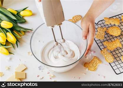 Cropped shot of woman using mixer machine making cookie icing for decoration. Woman holding glass bowl with sugar glaze and electric mixer. Easter holiday backing. Woman whisk cookie icing for decoration