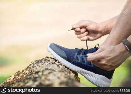 Cropped shot of young man runner tightening running shoe laces, getting ready for jogging exercise outdoors. Male jogger lacing his sneakers standing on forest path before morning run.
