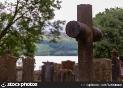 Cross and tombstones in cemetery with lake in background, in Luss parish church yard in Scotland