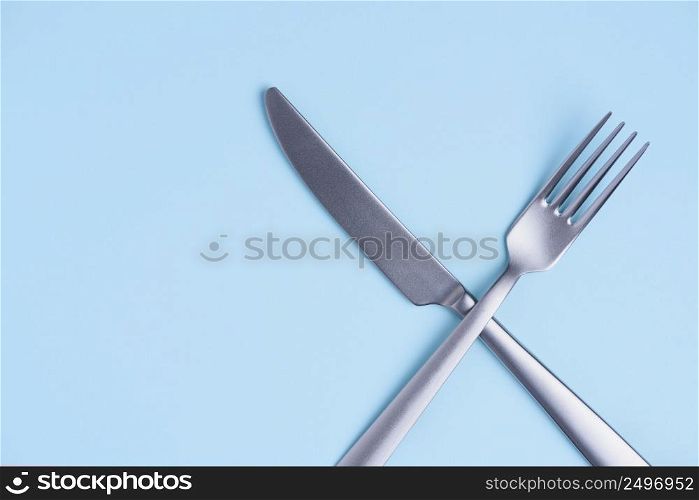 Crossed silver metal knife and fork sign on trendy blue pastel background with side copy space