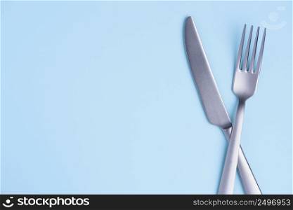 Crossed steel knife and fork on tredny soft blue paper background with side copy space