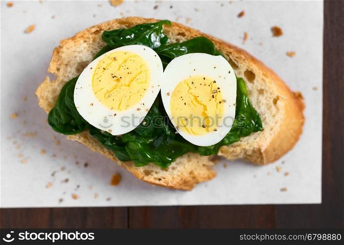 Crostini roasted bread slice with cooked spinach leaves and hard boiled quail eggs seasoned with black pepper, photographed overhead with natural light (Selective Focus, Focus on the top of the egg halves)