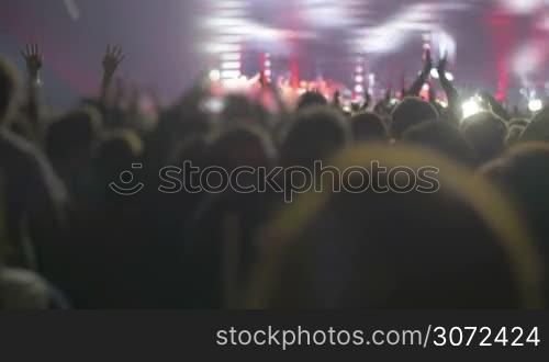 Crowd applauding during rock or pop concert in front of illuminated colored stage with huge screen showing singer