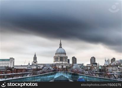 Crowds of locals and tourists cross the river Thames in London over a footbridge, with the dome of St. Paul&rsquo;s Cathedral under menacing rain clouds. Long exposure.