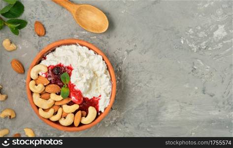 Crumbly fresh cottage cheese with sour cream, cranberry jam and nuts in traditional clay bowl, next to wooden spoon. Neutral gray background. Healthy breakfast made from curd, top view with copy space
