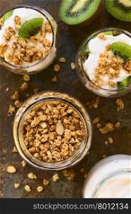 Crunchy almond and oatmeal granola in jar with yogurt kiwi granola parfait in glasses on the side, photographed overhead on slate with natural light (Selective Focus, Focus on the top of the granola and the parfaits)