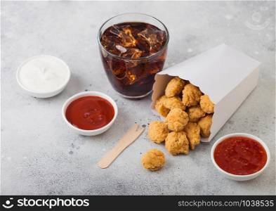 Crunchy southern chicken popcorn bites in white paper container for fast food meals with ketchup and glass of cola on light stone background.