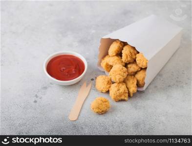 Crunchy southern chicken popcorn bites in white paper container for fast food meals with ketchup on light stone background.