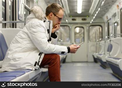 Crying young man looking at phone gets bad news, receiving message/sms, covering mouth with hand, sitting on seat in empty subway train, railway carriage on background. Problem in relationships.