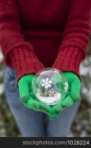 Crystal christmas ball with house and snow inside. Hands with green gloves hold transparent ball.