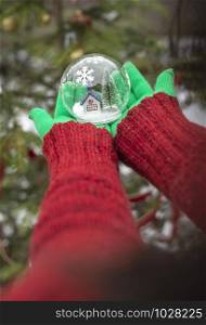 Crystal christmas ball with house and snow inside. Hands with green gloves hold transparent ball.