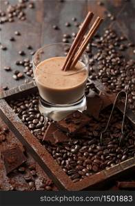 Crystal glass of Irish cream baileys liqueur with cinnamon, coffee beans and powder with dark chocolate in wooden tray on dark wood background. Top view