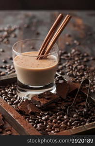 Crystal glass of Irish cream baileys liqueur with cinnamon, coffee beans and powder with dark chocolate in wooden tray on dark wood background. Top view