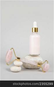 Crystal rose quartz facial roller and anti-aging collagen, serum in glass bottle on stones, grey background. Facial massage for natural lifting, Beauty concept Front view.. Crystal rose quartz facial roller and anti-aging collagen, serum in glass bottle on stones, grey background. Facial massage for natural lifting, Beauty concept Front view