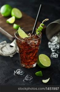 Cuba Libre cocktail in heavy crystal glass with ice and lime slice and peel, with straw and fresh limes with bar strainer on black table background.