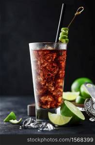 Cuba Libre cocktail in highball glass with ice and lime peel on bamboo stick with straw and fresh limes on black table background.
