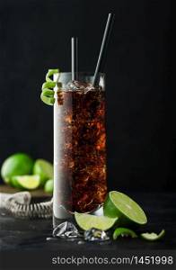 Cuba Libre cocktail in highball glass with ice and lime peel with straw and fresh limes and strainer on black table background.