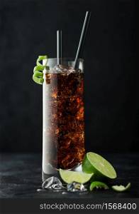 Cuba Libre cocktail in highball glass with ice and lime peel with straw and fresh limes on black table background.