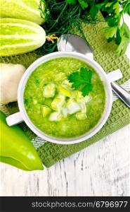 Cucumber soup with green peppers and garlic in a white bowl on a napkin, parsley on a background of wooden boards on top