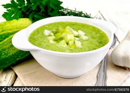 Cucumber soup with green peppers and garlic in a white bowl on a napkin, spoon, parsley on a wooden boards background