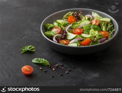 Cucumbers and tomatoes, red onion and spinach mix in fresh vegetables salad in grey bowl plate on dark background with pepper.