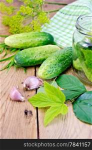 Cucumbers in a glass jar and on the table, garlic, tarragon, dill, leaves of cherry and currants on a wooden boards background