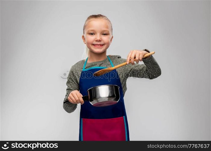 culinary and profession concept - happy smiling little girl in apron with saucepan and wooden spoon cooking food over grey background. little girl in apron with saucepan cooking food