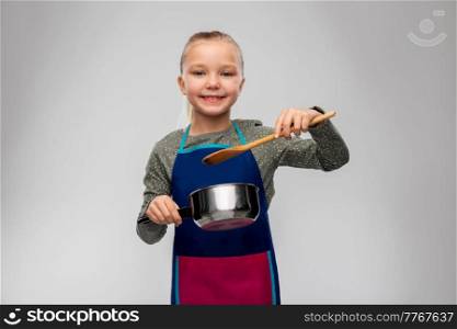 culinary and profession concept - happy smiling little girl in apron with saucepan and wooden spoon cooking food over grey background. little girl in apron with saucepan cooking food
