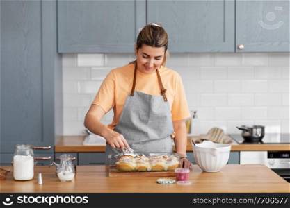 culinary, bake and cooking food concept - happy smiling young woman adding buttercream topping to baked roll buns in baking dish on kitchen at home. woman cooking food and baking buns at home kitchen
