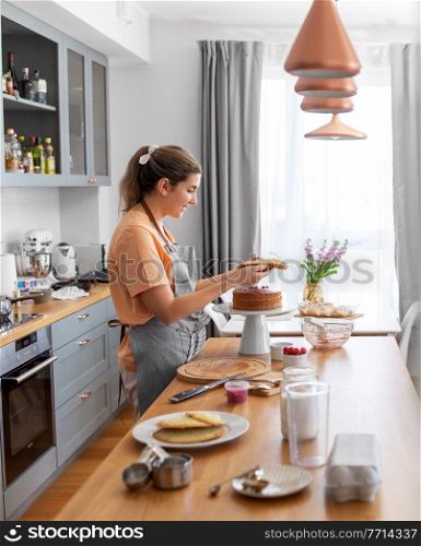 culinary, baking and cooking food concept - happy smiling young woman making layer cake on kitchen at home. woman cooking food and baking on kitchen at home