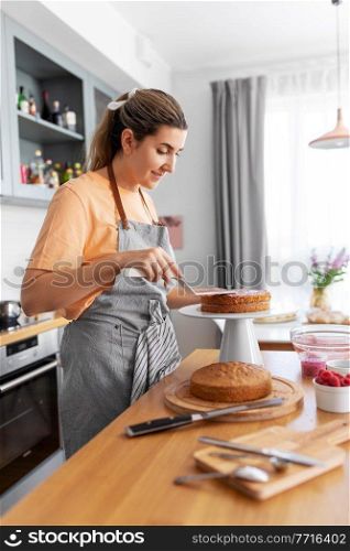 culinary, baking and cooking food concept - happy smiling young woman making layer cake on kitchen at home. woman cooking food and baking on kitchen at home