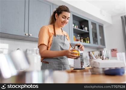 culinary, baking and people concept - happy smiling young woman cooking food on kitchen at home and grating lemon peel on grater. happy young woman cooking food on kitchen at home