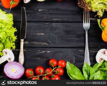 Culinary black frame background with fresh farm vegetables, yellow paprika pepper, basil, cherry tomatoes, mushrooms, red onion, lime, lettuce. Food top view. Knife and fork in the middle