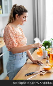 culinary, drinks and people concept - happy smiling young woman making orange cocktail at home kitchen. woman making cocktail drinks at home kitchen