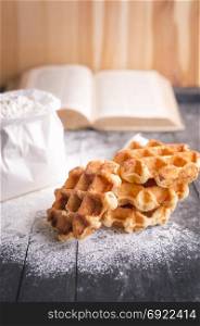 Culinary theme image with a bunch of delicious, freshly baked belgian waffles surrounded by wheat flour and an open cookbook, on a wooden table.