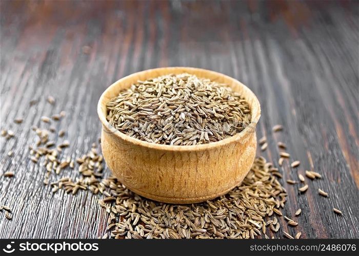 Cumin seeds in a bowl and on a table against dark wooden board background