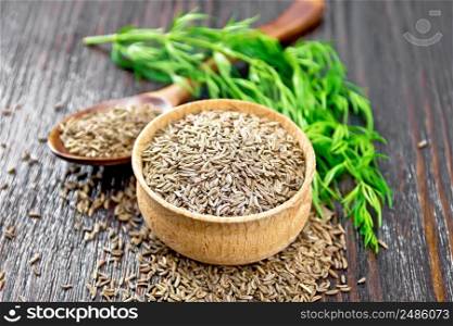 Cumin seeds in a bowl, spoon and on the table, a green sprig of caraway on dark wooden board background