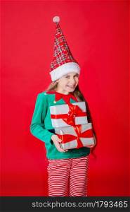 Cunning happy attractive girl with gifts in a costume of Santa Claus helper elf on a bright red bright color background. Portrait of a beautiful elven baby. Copy space.. Happy attractive girl with gifts in a costume of Santa Claus helper elf on a bright red bright color background.