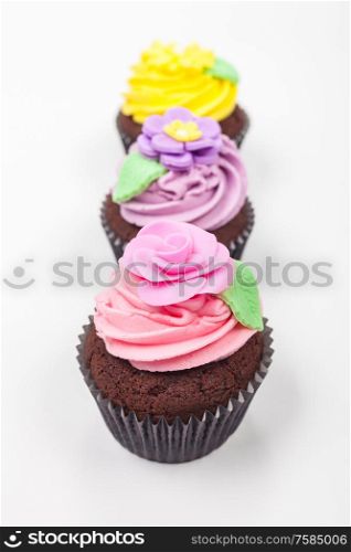 Cup cakes or cupcakes with icing or frosting, pink, purple and yellow with green leaves, rose and floral decorations photographed on a white background