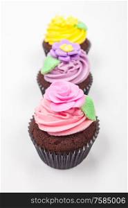 Cup cakes or cupcakes with icing or frosting, pink, purple and yellow with green leaves, rose and floral decorations photographed on a white background