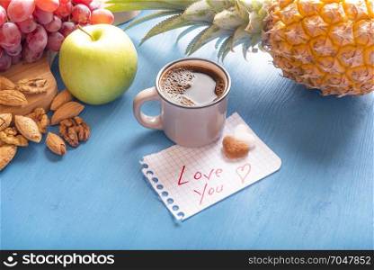 Cup of aromatic coffee, heart shaped sugar pieces and a love you message on a math paper, surrounded by fresh fruits and nuts, on a blue wooden table.