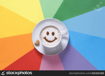 Cup of cappuccino with a smiley face, on the milk froth from cocoa powder and a heart-shaped sugar near it, on a paper background in rainbow colors.