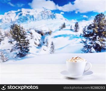 cup of cappuccino with whipped cream on wooden table over winter landscape