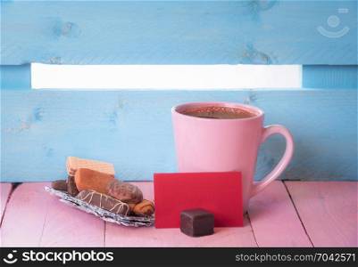 Cup of coffee near a pile of sweets on a tree bark and a blank red paper note, on a pink wooden table and a blue fence in the background.