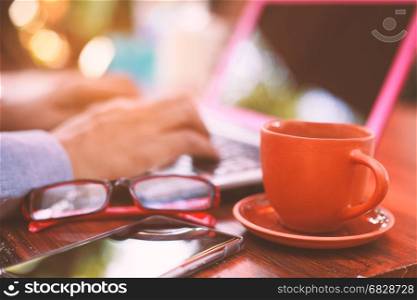 Cup of coffee on the wood with smartphone , glasses and hand writting at laptop.