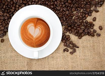 Cup of coffee with heart shape in foam and beans on hessian background