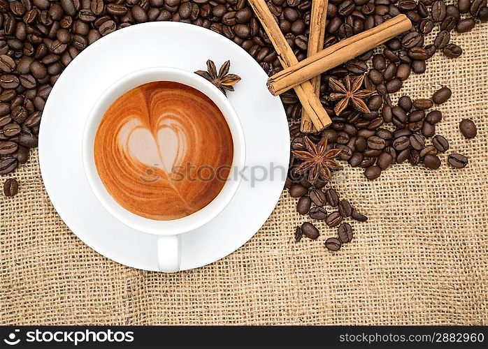 Cup of coffee with heart shape in foam with coffee beans and cinnamon sticks and star anise on hessian background