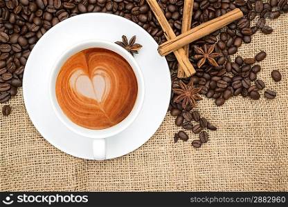 Cup of coffee with heart shape in foam with coffee beans and cinnamon sticks and star anise on hessian background